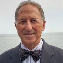 Hombre con cabello corto gris, vestido con un traje gris y una corbata azul oscuro, sonriendo a la cámara. 