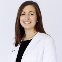 Mujer sonriente con cabello castaño oscuro y bata blanca con camisa negra. 