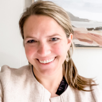 Mujer rubia sonriente con el pelo recogido en una coleta y chaqueta beige. 