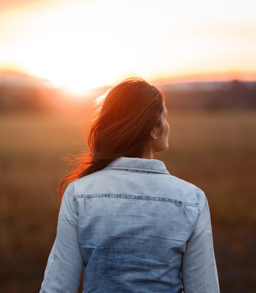 Imagen decorativa de una persona con el pelo largo de espaldas, mirando hacia un amanecer. 